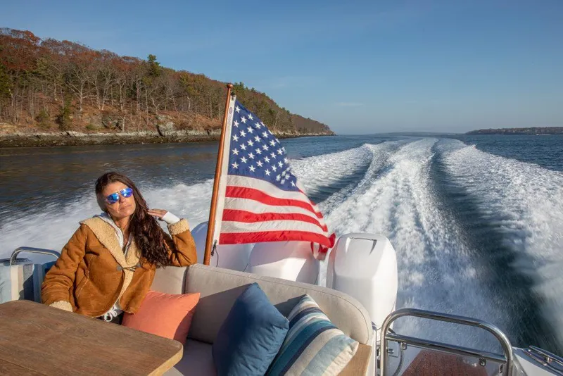 Slide: The Image of Woman relaxing on a 2025 Back Cove 390 boat with American flag, scenic coastal view. - 32