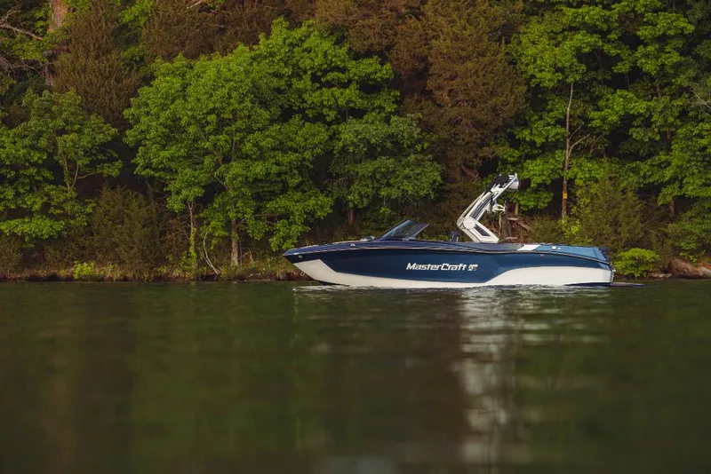 Slide: The Image of 2023 MasterCraft XT22 T boat on a calm lake with a forested background. - 0