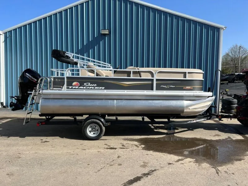 The Image of 2023 Sun Tracker PARTY BARGE® 18 DLX pontoon boat on trailer outside blue building. - 0