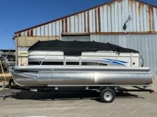 Slide: The Image of 2023 Sweetwater 2086C pontoon boat on trailer, parked near a rustic metal building. - 0