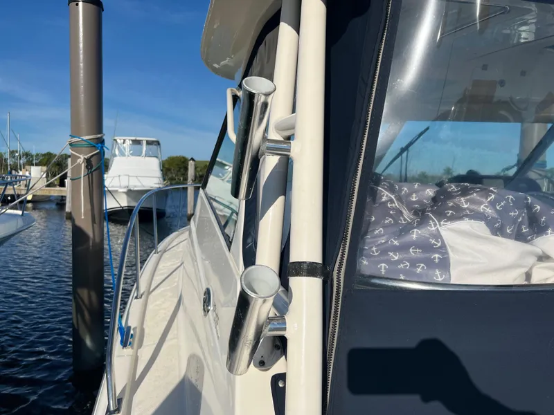 Slide: The Image of 2014 Pursuit 345 OS boat docked, featuring rod holders and cabin window. - 49