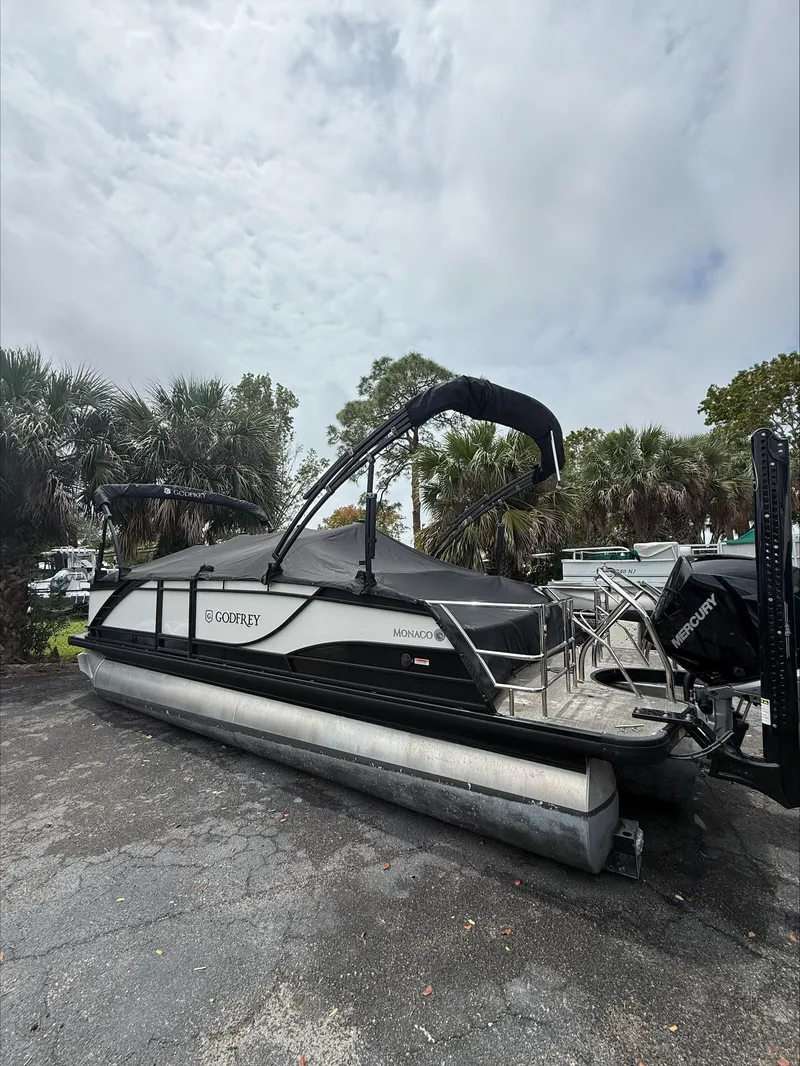 Slide: The Image of 2023 MONACO 235 DFL pontoon boat with canopy, parked outdoors under cloudy sky. - 1