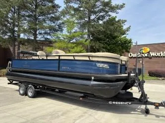 Slide: The Image of 2023 Regency 250 DL3 pontoon boat on trailer, parked outdoors near trees and building. - 1