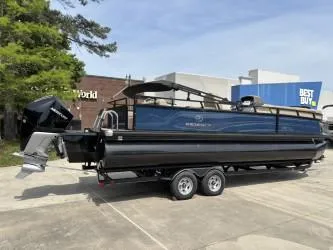 Slide: The Image of 2023 Regency 250 DL3 pontoon boat on trailer, parked near retail stores. - 2