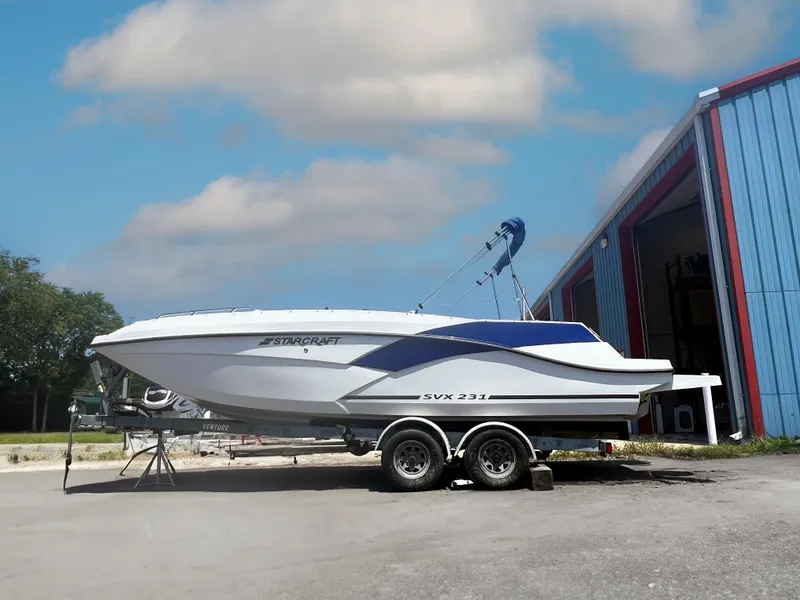 Slide: The Image of 2023 Starcraft SVX 231 OB DH boat on trailer, parked outside a blue building. - 1