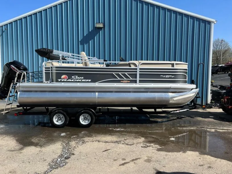 The Image of 2023 Sun Tracker PARTY BARGE® 20 DLX pontoon boat on a trailer outside a blue building. - 0