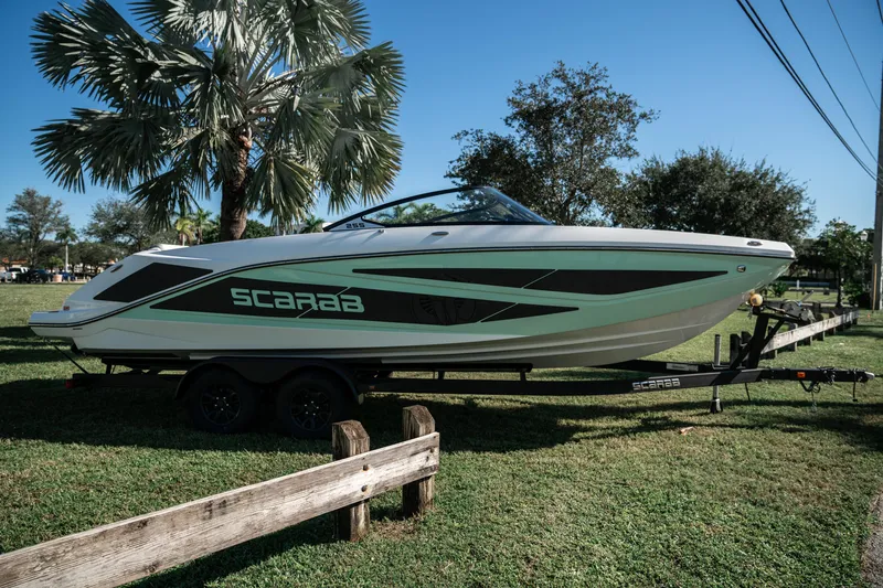 Slide: The Image of 2022 Scarab 255 boat on a trailer, parked on grass with trees in the background. - 2