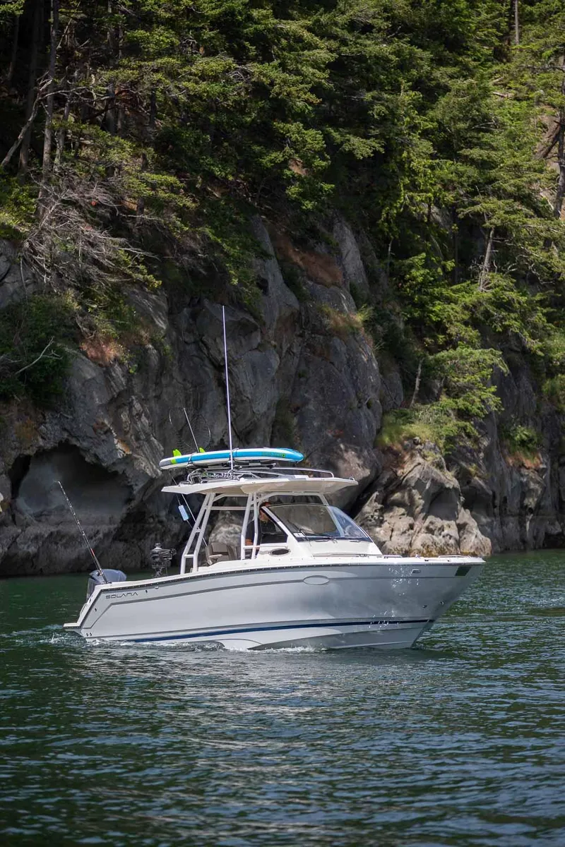 Slide: The Image of 2023 Solara S-250 CW boat cruising near rocky shoreline with lush greenery. - 2