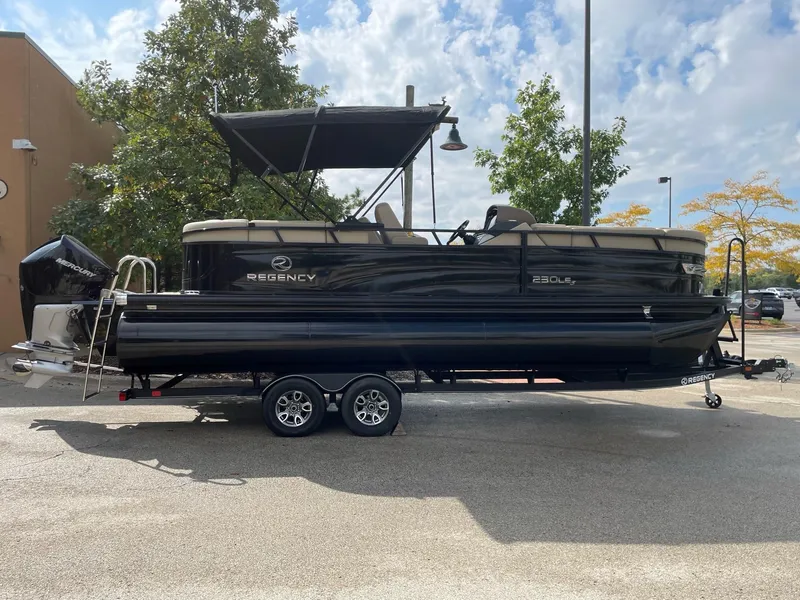 Slide: The Image of 2023 Regency 230 LE3 pontoon boat on a trailer, black exterior, parked outdoors. - 0