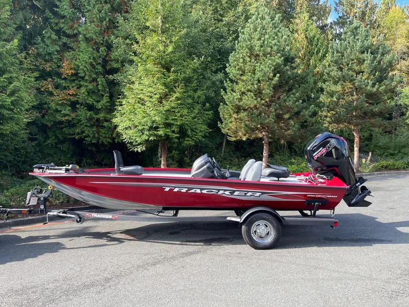 The Image of 2023 Tracker Pro Team 190 TX boat on trailer, parked near lush green trees. - 0