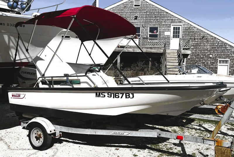 Slide: The Image of 2002 Boston Whaler 130 Sport boat with red canopy on trailer, parked near a wooden building. - 2