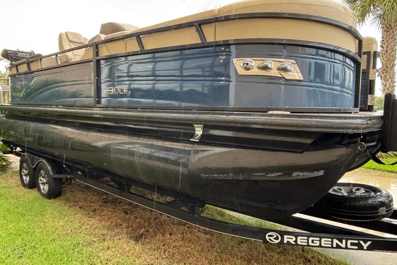 The Image of 2023 Regency 230 LE3 pontoon boat on trailer, side view, blue and black design. - 0