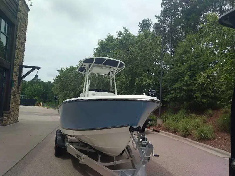 Slide: The Image of 2023 Mako 214 CC boat on a trailer, parked outdoors near trees and a building. - 2