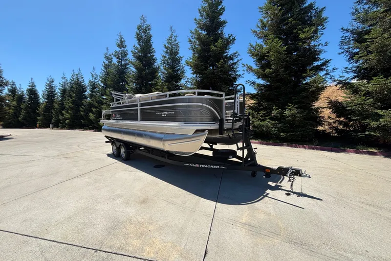 Slide: The Image of 2024 Sun Tracker Fishin' Barge 20 DLX pontoon boat on trailer, surrounded by trees. - 2