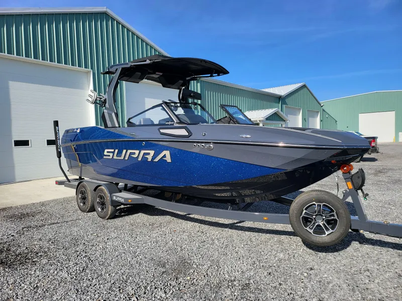 Slide: The Image of 2024 Supra SL boat on trailer, parked near green warehouses under clear blue sky. - 1