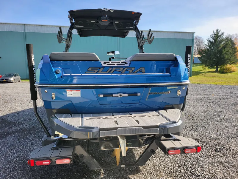 Slide: The Image of 2024 Supra SL boat, blue exterior, rear view, parked on gravel. - 2