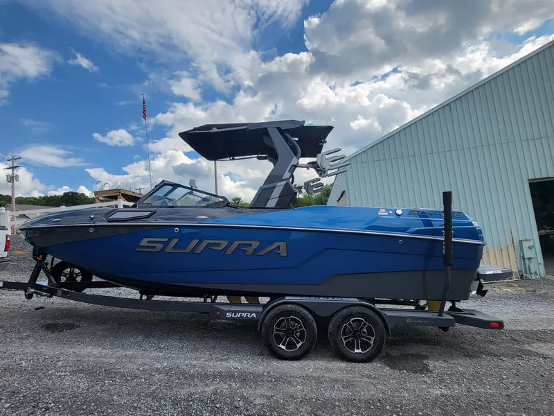 Slide: The Image of 2024 Supra SL boat on trailer, blue exterior, parked outdoors under cloudy sky. - 23