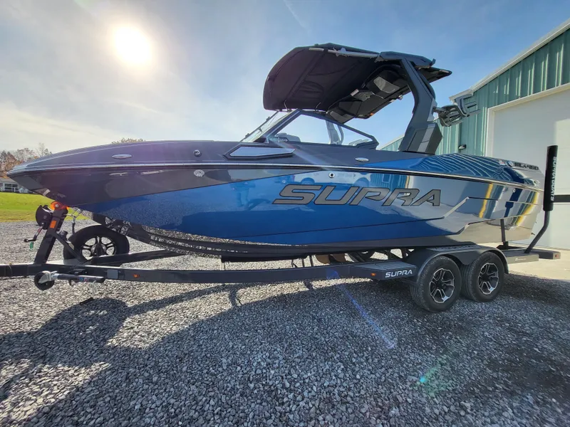 Slide: The Image of 2024 Supra SL boat on trailer, blue exterior, parked outdoors in sunlight. - 29
