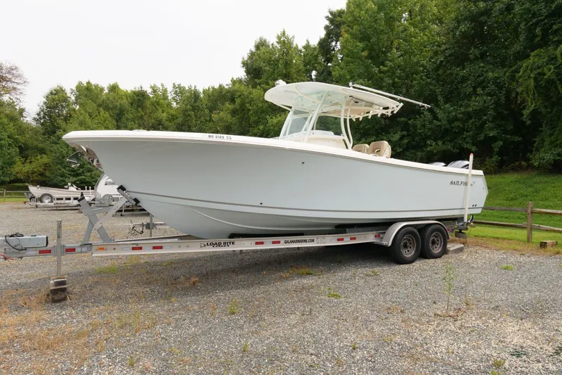 The Image of 2018 Sailfish 290 CC boat on a trailer in a gravel lot with trees in the background. - 0