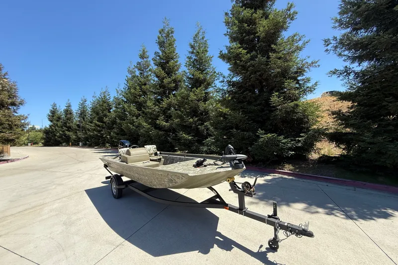 Slide: The Image of 2024 Tracker Grizzly 1754 SC boat on trailer, parked outdoors near trees. - 2