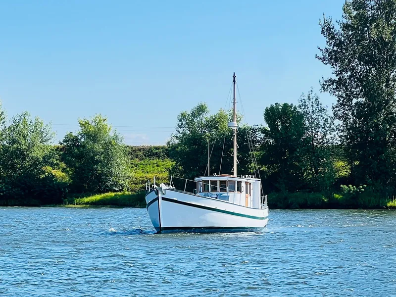 Slide: The Image of 1955 Colin Archer 42 boat sailing on a calm river with lush green trees in the background. - 43