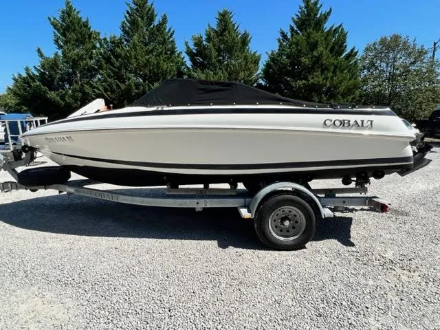 Slide: The Image of 2001 Cobalt 190 boat on trailer, covered, parked on gravel lot with trees in background. - 23