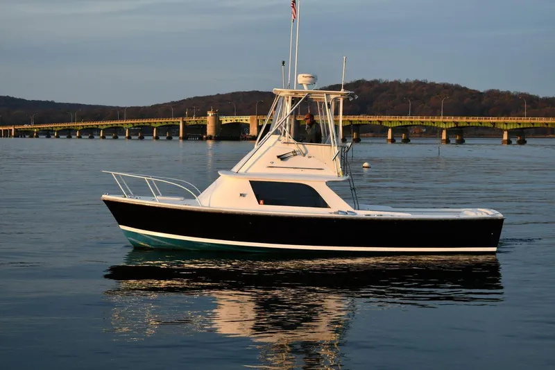 The Image of 1965 Bertram Sportfisher boat on calm water near a bridge at sunset. - 1