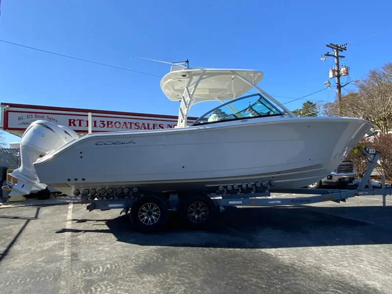 Slide: The Image of 2025 Cobia 280 Dual Console boat on trailer at dealership under clear blue sky. - 2