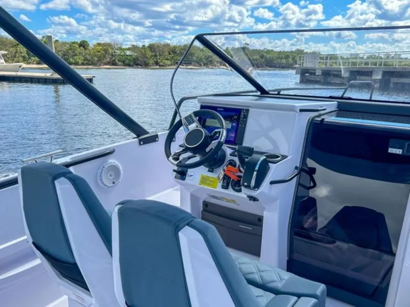Slide: The Image of 2024 Axopar 25 Cross Top boat interior with steering wheel and dashboard, docked by a scenic lake. - 14
