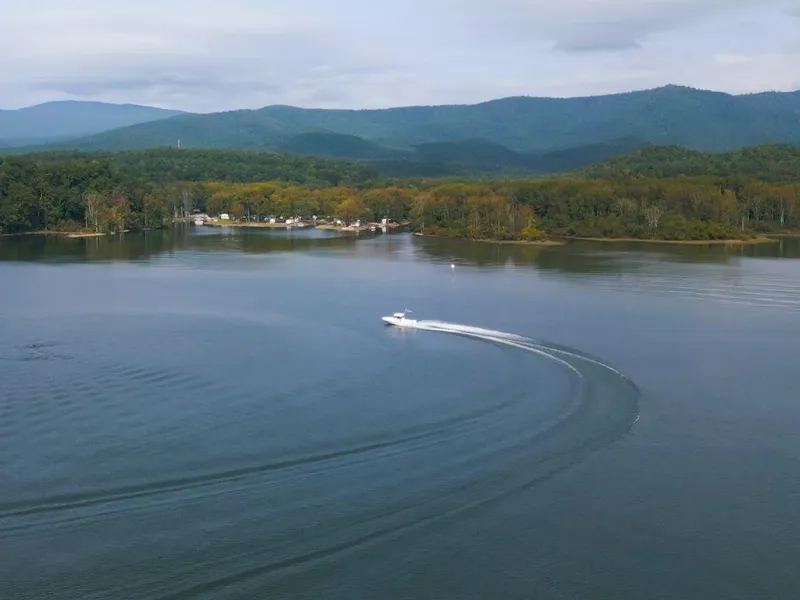 Slide: The Image of 2024 Sportsman Masters 267OE Bay boat cruising on a serene lake with forested hills. - 7