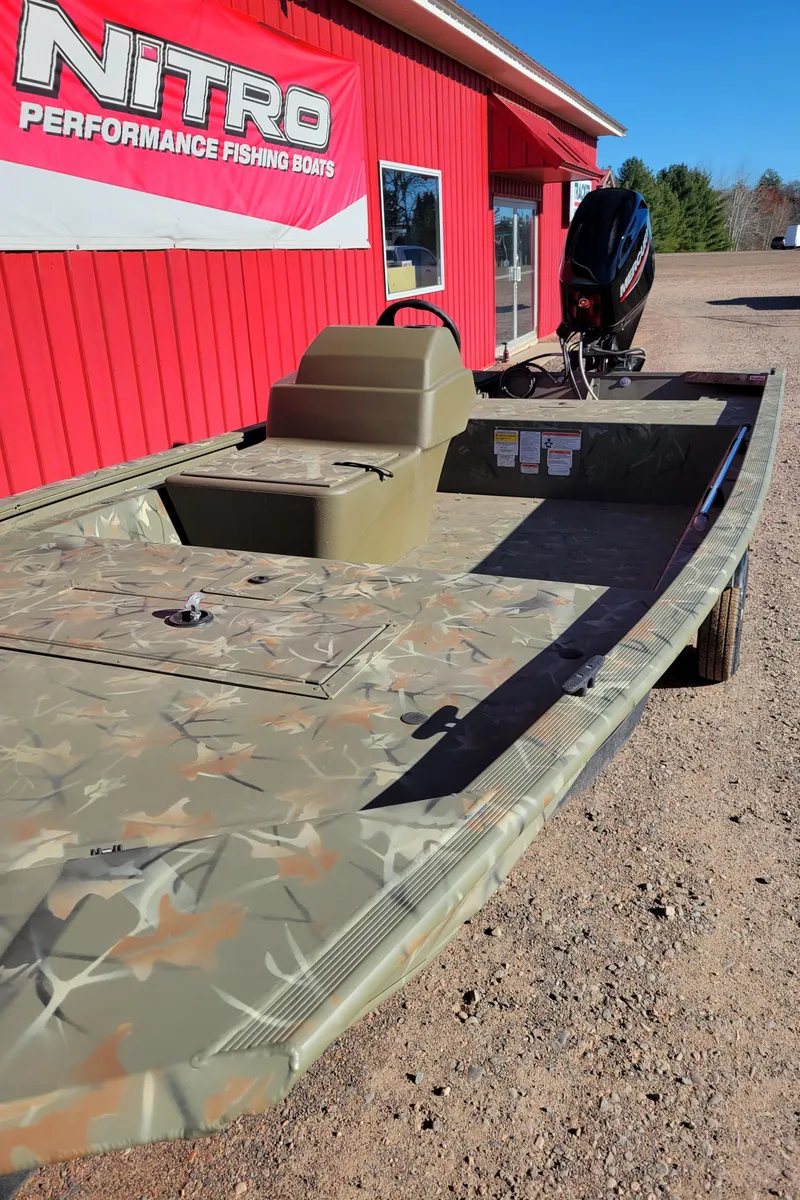 Slide: The Image of 2024 Tracker Grizzly 1648 SC fishing boat in camouflage, parked outside a red building. - 6