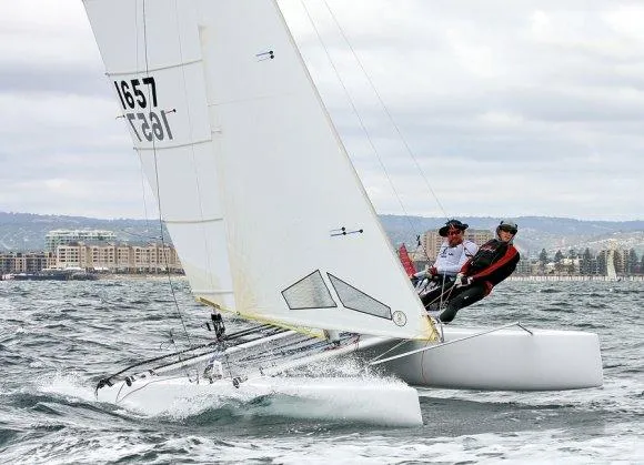 The Image of Sailors navigating a 1989 Nacra 5.8 catamaran on choppy waters near a coastal city. - 1