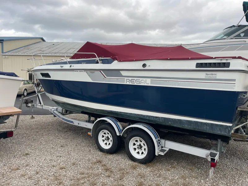 Slide: The Image of 1988 Regal boat on trailer with maroon cover, parked on gravel. - 1