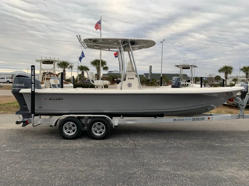 Slide: The Image of 2025 Bulls Bay 2400 boat on trailer, parked outdoors with flags and palm trees in background. - 5