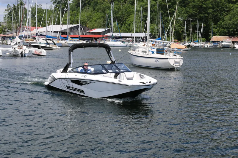 Slide: The Image of 2024 Scarab 215 Wake LE boat cruising in a marina with sailboats in the background. - 2