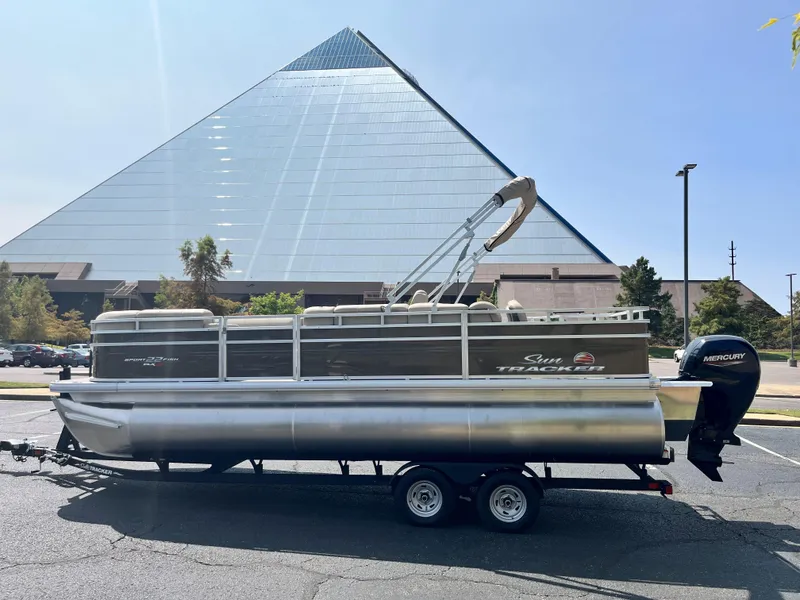The Image of 2024 Sun Tracker SportFish 22 XP3 pontoon boat on trailer, parked near a large glass pyramid. - 0
