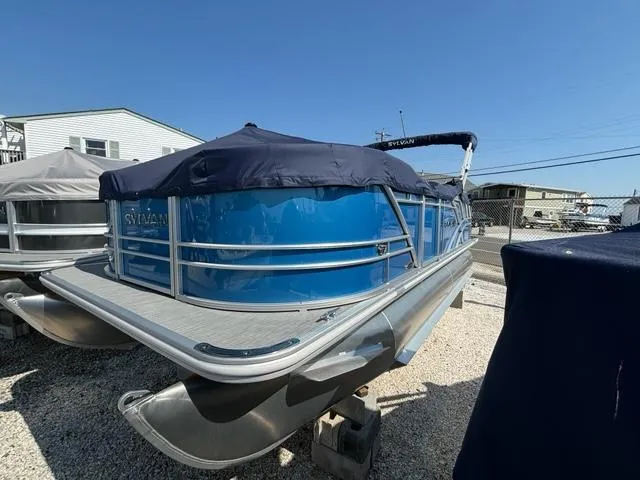 Slide: The Image of 2024 Sylvan Mirage 8520 Cruise pontoon boat with blue cover, parked on gravel. - 2