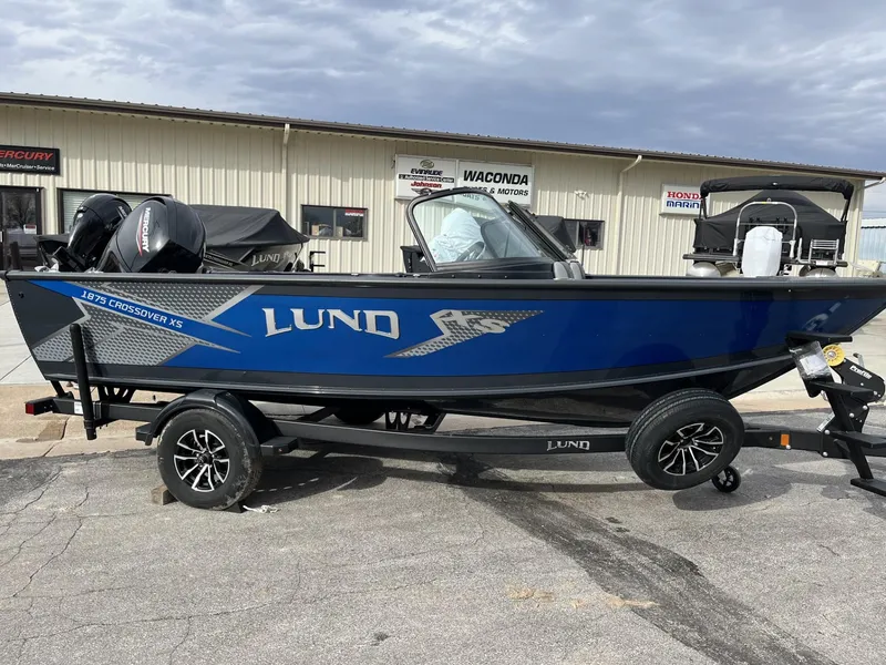 Slide: The Image of 2025 Lund 1875 Crossover XS boat on trailer, blue and black, parked outside dealership. - 2