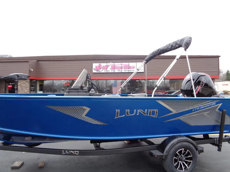 Slide: The Image of 2024 Lund 1775 Adventure SS boat in blue, parked outside a dealership. - 2