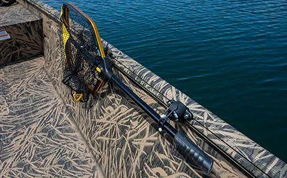 Slide: The Image of 2024 Crestliner 1660 Retriever SC boat with camouflage design and fishing net by the water. - 11