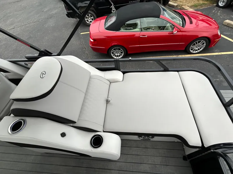 Slide: The Image of 2024 Avalon LSZ 2585 RL boat lounge seating with cup holders, parked red convertible in background. - 21