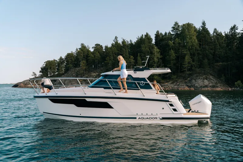 Slide: The Image of 2024 Aquador 250 HT boat cruising on a serene lake with lush forest backdrop. - 13