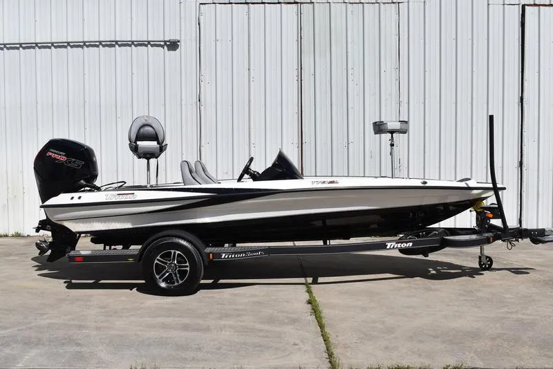 Slide: The Image of 2024 Triton 179 TrX boat on trailer, parked outdoors against a metal wall. - 2