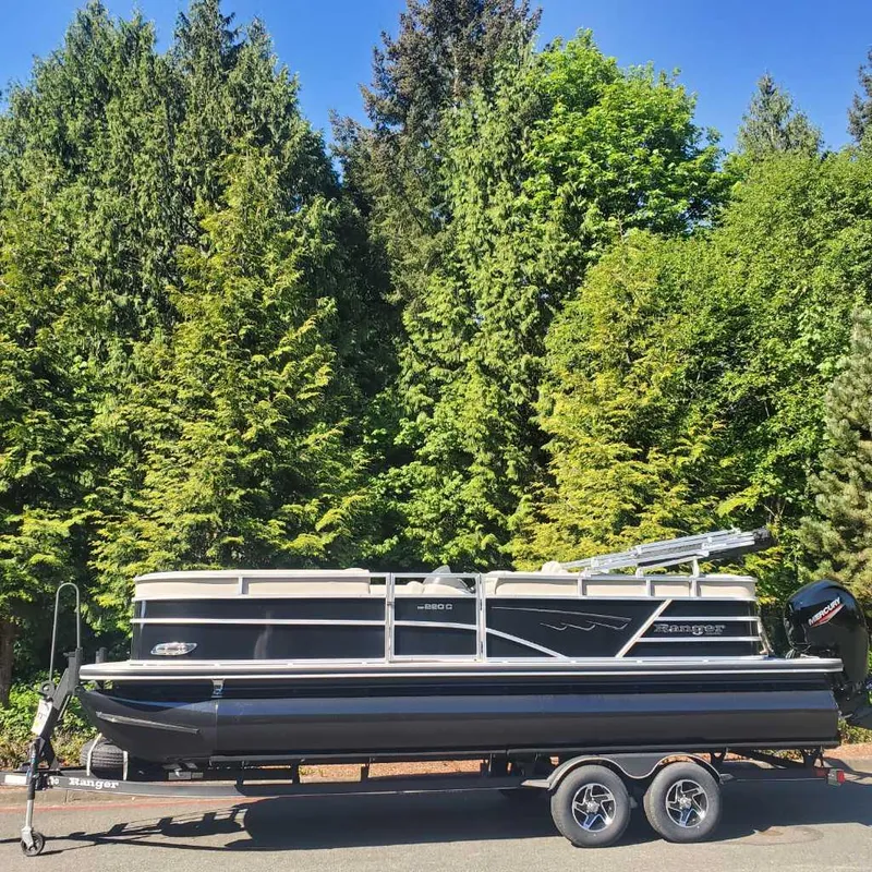 The Image of 2024 Ranger Reata 220C pontoon boat on trailer, surrounded by lush green trees. - 0