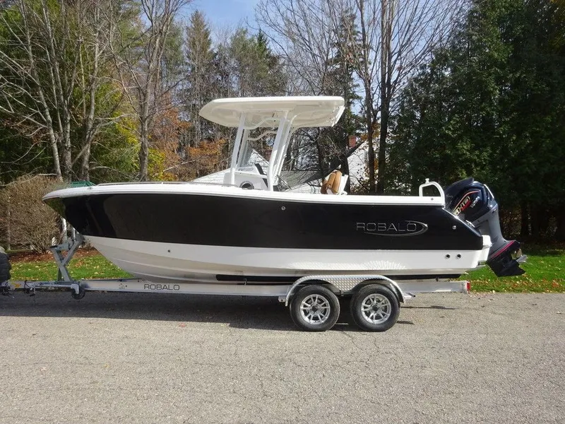 Slide: The Image of 2024 Robalo R230 boat on trailer, parked outdoors, surrounded by trees. - 0