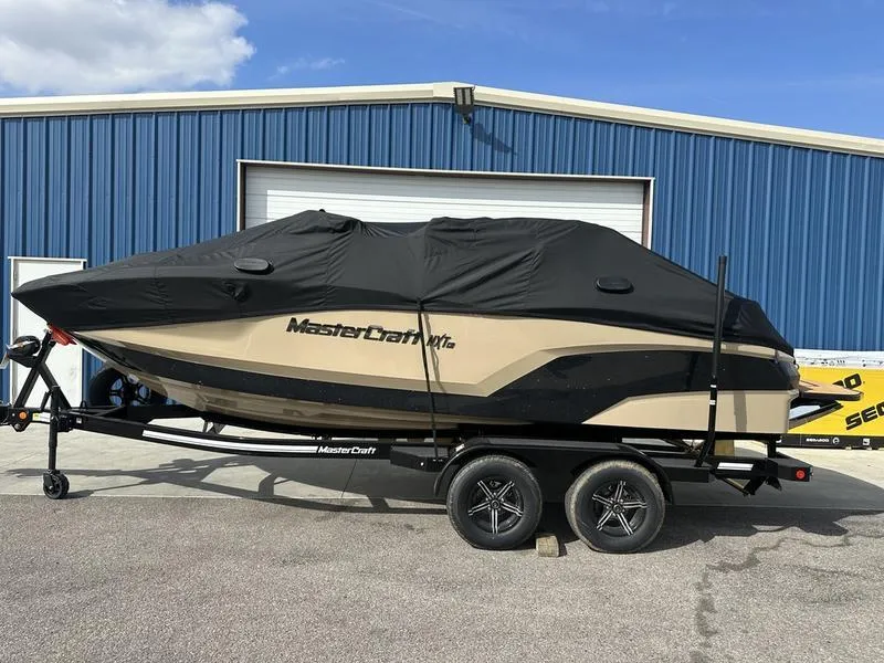 Slide: The Image of 2024 MasterCraft NXT21 boat on trailer with black cover, parked outside blue building. - 2