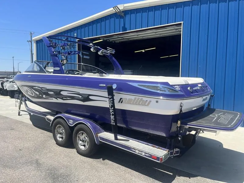 Slide: The Image of 2007 Malibu 247 LSV boat on trailer, blue and white, outside blue building. - 2