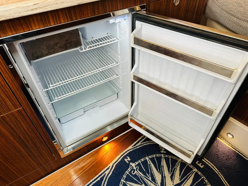 Slide: The Image of Open refrigerator inside 2017 Pursuit OS 355 boat, featuring wooden cabinetry and nautical-themed rug. - 49