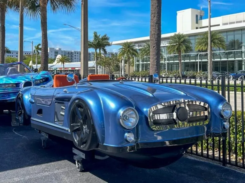 Slide: The Image of 2023 Watersports Car Series M, blue, parked outdoors with palm trees and modern buildings in the background. - 1