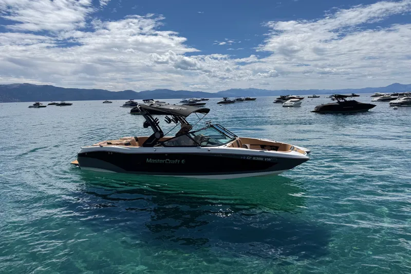 The Image of 2024 MasterCraft X26 boat on a serene lake with distant mountains and scattered clouds. - 0